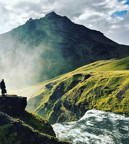 A person, black silhouette standin on a rock observing a river and a waterfall. Green hill and black mountain in background